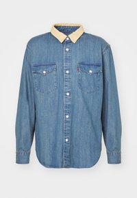 Camicia in denim blu con maniche lunghe, colletto giallo, chiusura frontale con bottoni e due taschini sul petto. Presenta un orlo curvo e dettagli di cucitura.