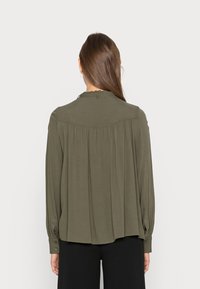 Blusa de mangas longas verde-oliva com um design traseiro plissado e punhos com botão. Tecido suave e leve com colarinho redondo.
