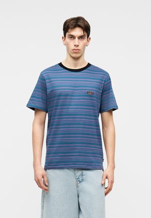 Mladi moški v modro-vijolični črtasti majici s črnim ovratnikom in svetlo modrih jeans kratkih hlačah stoji pred enobarvnim ozadjem.