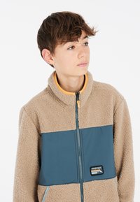Chaqueta de forro polar beige con un panel azul en el pecho, cuello alto, cremallera completa y un parche de logo decorativo en el frente.