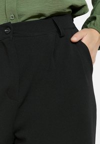 Pantaloni neri su misura realizzati in un tessuto liscio, dotati di tasche laterali, chiusura con bottone e vita aderente, abbinati a una maglietta verde.