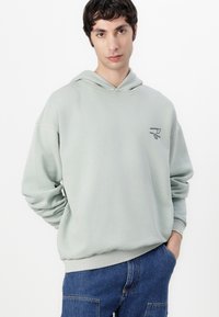 Hellgrüner Hoodie mit Kordelzug, ausgestattet mit einem kleinen schwarzen Logo auf der Brust, aus weichem Material gefertigt, mit gerippten Bündchen und Saum.