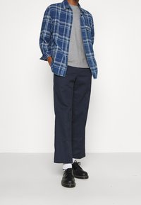 Modrá kostkovaná flanelová košile přes šedé tričko s kulatým výstřihem, v kombinaci s tmavě modrými kalhotami a černými šněrovacími botami. Viditelné bílé ponožky.