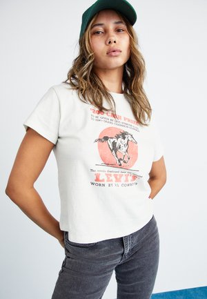 Hvid kortærmet t-shirt med et grafik af en løbende hest og rød tekst, parret med mørkegrå jeans og en grøn kasket.
