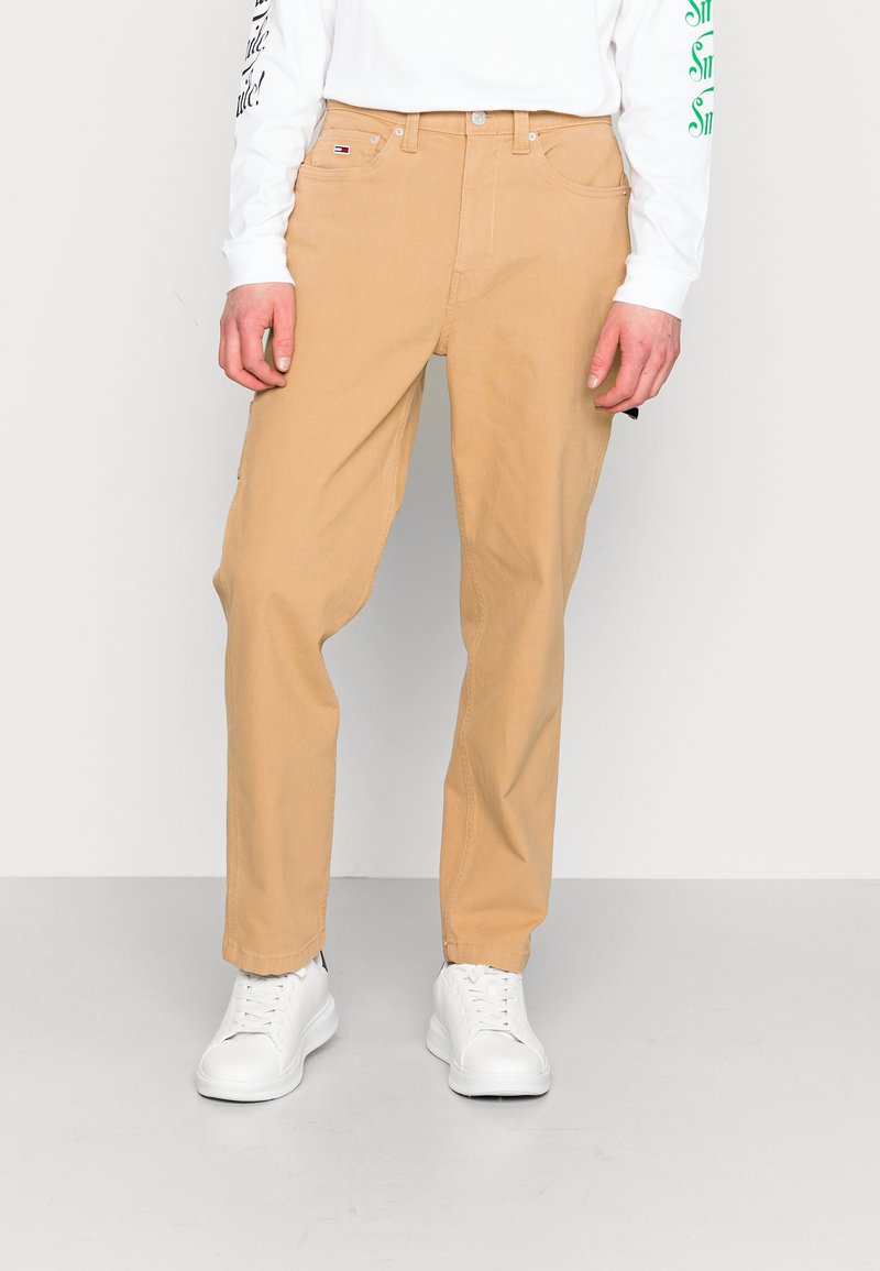 Chinos en coton beige avec une coupe décontractée, dotés de poches avant, d'une fermeture à bouton et d'un branding subtil sur la taille. Associés à des baskets blanches.