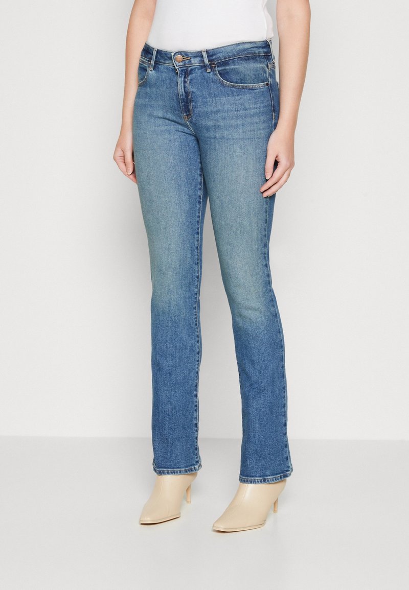 Femme portant un jean bleu taille mi-haute à jambes droites et des bottines beiges à talons, debout devant un fond blanc uni.