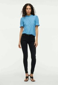 Kvinne med krøllete hår iført en blå blonderkortermet topp, sorte skinny jeans og svarte høye sandaler, står mot en ensfarget bakgrunn.