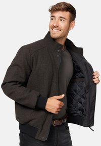 Smeđa teksturirana jakna s uzdignutim ovratnikom, zatvaranjem na patentni zatvarač i elastičnim manžetama. Unutarnja podstava je crna i quilted. Model prikazuje jaknu djelomično otvorenu.