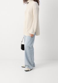 Crèmekleurige gebreide coltrui, comfortabele pasvorm; lichtblauwe wijde jeans; zwarte handtas; lichtgekleurde platte schoenen met een spitse neus.