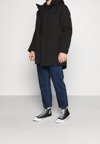 Mand iført sort parka, blå opstammede jeans og sorte høj-top sneakers står mod en ensfarvet hvid baggrund.