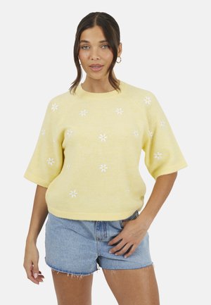 Donna con capelli castani, indossa un maglione giallo a maniche corte con fiori bianchi e pantaloncini di jeans, in piedi con una mano sui fianchi.