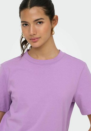 Ung kvinde med mørkt hår samlet bagud, iført gyldne øreringe og en lavendel kortærmet t-shirt med rund hals, positurerer mod en ensfarvet baggrund.