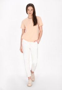 ferskenfarvet bomulds T-shirt med rulleærmer, hvide tapered bukser og beige sneakers. Modellen står med den ene hånd i lommen, neutral baggrund.