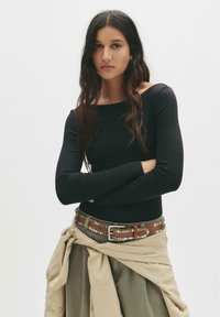 Långärmad svart topp, figurnära design. Modellen bär ett brunt bälte med silvernitar över ett beige plagg knutet i midjan. Enkla vita bakgrund.