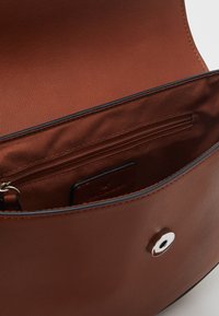 Borsa a tracolla in pelle marrone aperta che rivela un interno liscio e bronzo con una tasca con zip e un'etichetta di branding visibile.