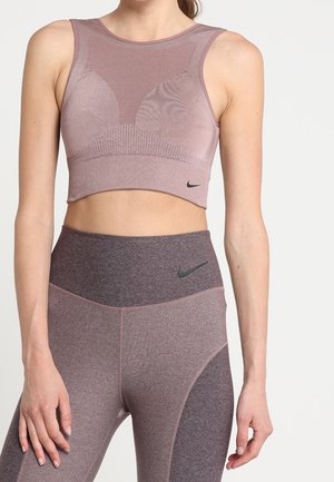 Vrouw draagt een mauve naadloze cropped top en hoge taille legging met Nike-logo's, staand tegen een effen lichte achtergrond.