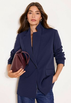 Nainen, jolla on yllään tummansininen bleiseri ja farkut, pitää tumman marjanväristä käsilaukkua toisessa kädessään ja toinen käsi on taskussa, yksinkertaisen taustan edessä.