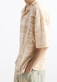 Korte mouwen beige shirt met abstracte tan patronen; lichtgewicht stof; met een kraag en knoopdetails. Gedragen met lichtblauwe jeans.