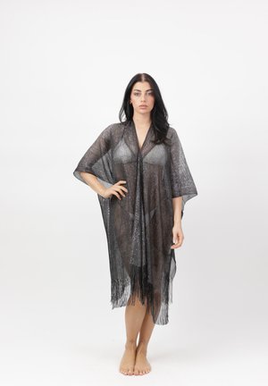 Kvinde stående barfodet med hånden på hoften iført et gennemsigtigt sort frynset sjal over en sølvfarvet bikini mod en ensfarvet hvid baggrund.