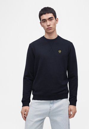 Unge mand med kort sort hår iført en sort crewneck sweatshirt med et lille logo på brystet og lyseblå jeans.