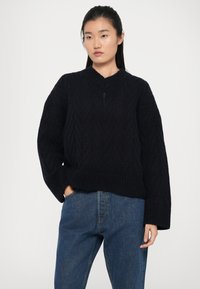 Maglione nero lavorato a maglia con un motivo a zigzag testurizzato, colletto alto e maniche ampie, abbinato a jeans blue denim.