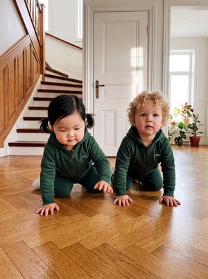 Deux tout-petits vêtus de tenues vertes assorties rampent sur un plancher en bois dans une maison lumineuse, à côté d’un escalier.