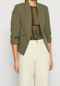 Zielona oliwkowa marynarka dopasowana, z kołnierzem stojącym, przednią kieszenią na zamek oraz podwiniętymi rękawami, zestawiona z ciemnym topem w tie-dye i jasnymi spodniami.