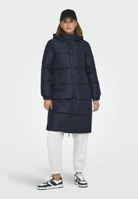 Manteau long matelassé bleu marine avec un grand col et une capuche, présentant une fermeture éclair à l'avant, des poches latérales et des poignets élastiques. Porté sur un pantalon de survêtement gris.
