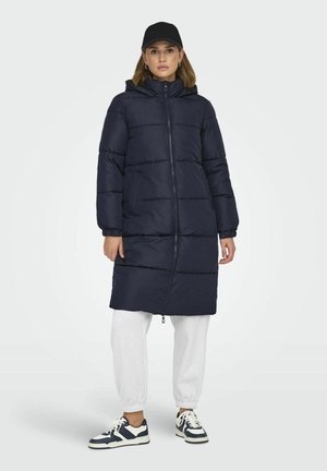 Manteau long matelassé bleu marine avec un grand col et une capuche, présentant une fermeture éclair à l'avant, des poches latérales et des poignets élastiques. Porté sur un pantalon de survêtement gris.