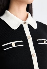 Cardigan en tricot noir avec un col blanc, comportant des boutons argentés et deux poches poitrine avec des accents blancs. Matière texturée, couleurs contrastées.