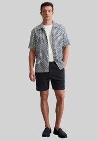 Camicia a maniche corte con bottoni in un motivo geometrico blu e bianco, abbinata a pantaloni corti scuri e mocassini neri, che offre una vestibilità comoda.