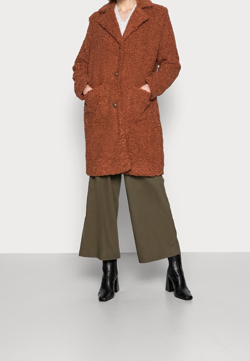 Personne portant un manteau teddy couleur rouille avec des revers, un pantalon large couleur olive et des bottines noires à talons, debout les mains dans les poches du manteau.
