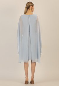 Robe bleu clair avec des manches en cape transparentes, tissu texturé et détail de fermeture éclair au dos. La longueur arrive au-dessus du genou avec une légère évasement.