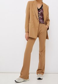 Un costume deux pièces en tissu beige comprend un blazer croisé à boutons dorés et un pantalon fuselé assorti, associé à un haut à motif floral.