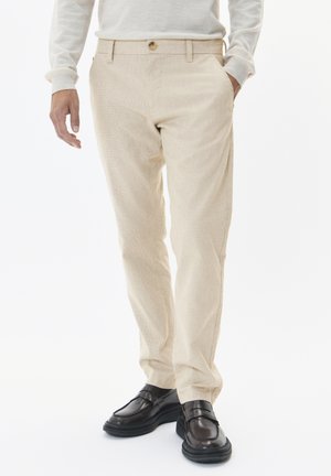 Pantalones beige texturizados con un corte ajustado, con cierre de botón en la parte delantera y bolsillos laterales, combinados con zapatos oscuros y pulidos.