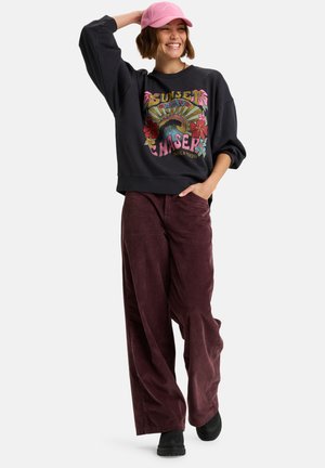 Donna che sorride con una mano sulla cuffia rosa, indossando una felpa nera con grafica, pantaloni di velluto a coste borgogna larghi e scarpe nere, su sfondo bianco.