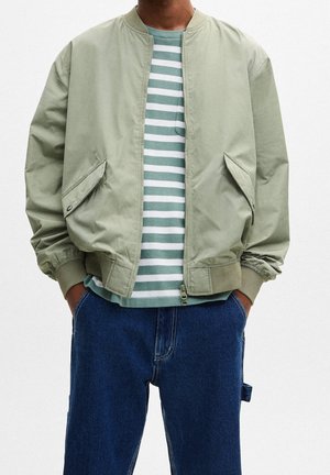 Personne portant une veste bomber vert clair sur une chemise rayée verte et blanche, un jean bleu et les mains dans les poches.