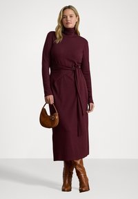 Abito lungo bordeaux con collo a lupetto e design avvolgente, caratterizzato da una cintura in tessuto e maniche lunghe. Abbinato a una borsa in suede marrone e stivali al ginocchio.
