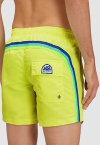 Costumi da bagno giallo neon con strisce laterali blu, verdi e verde chiaro. Include una tasca posteriore con un patch logo. Texture liscia.