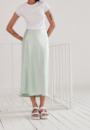 Woman with visible arm tattoos wearing a white crop top, light green midi skirt, and white platform sandals standing indoors.