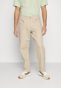 Pantalones de algodón beige con corte recto, que cuentan con bolsillos frontales y cierre de botón, combinados con zapatillas blancas y verdes.