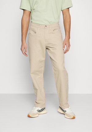 Pantalones de algodón beige con corte recto, que cuentan con bolsillos frontales y cierre de botón, combinados con zapatillas blancas y verdes.