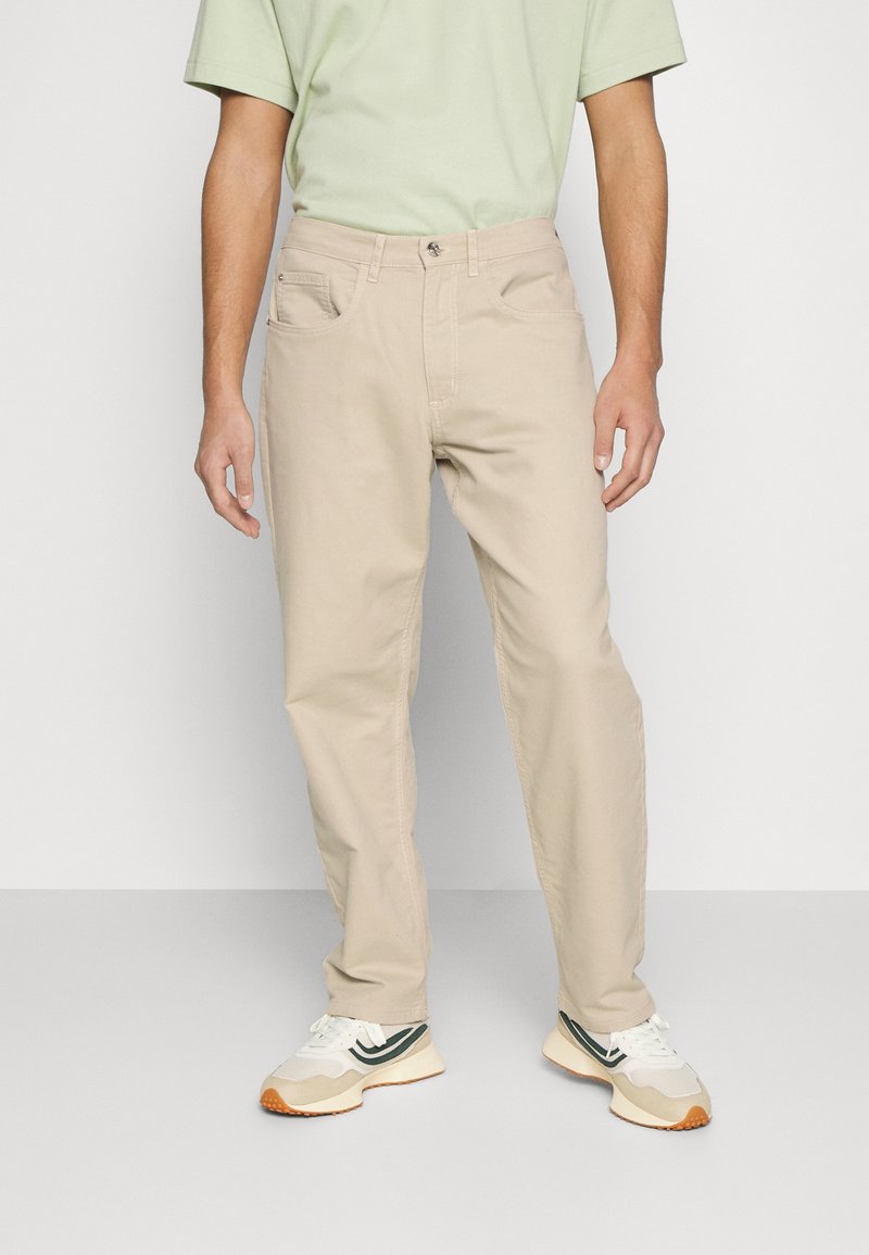 Pantalones de algodón beige con corte recto, que cuentan con bolsillos frontales y cierre de botón, combinados con zapatillas blancas y verdes.