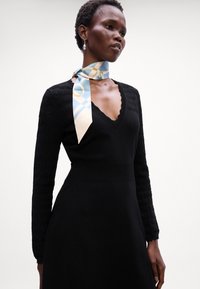 Abito nero lavorato a maglia con maniche lunghe a trama, scollatura a V scallop e una leggera sciarpa di seta blu con motivo legata attorno al collo.