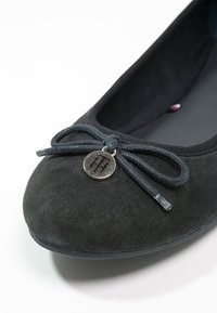 Ballerine en daim noir avec un nœud en tissu et un charm en métal rond ornée d'un logo embossé sur l'avant.