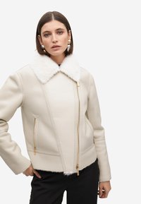Giacca di pelle color crema con colletto in pelliccia sintetica, cerniere dorate e tasche laterali. Presenta un design aderente e un tessuto testurizzato.