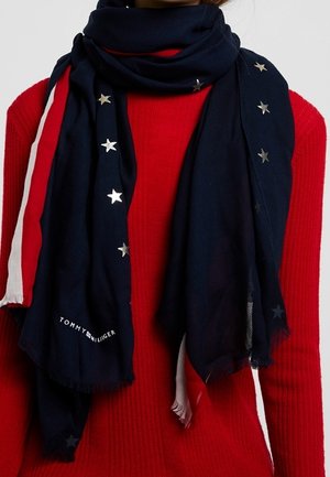 Écharpe bleu marine avec des étoiles argentées et le logo "Tommy Hilfiger" drapée sur un pull rouge côtelé avec des rayures blanches sur les manches.