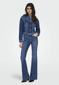 Chaqueta de mezclilla de color azul, con botones en el frente y dos bolsillos en el pecho. Combinada con unos jeans azules acampanados y unas botas negras hasta el tobillo. Textura suave.