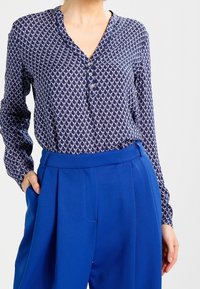 Blusa a fantasia navale con scollo a V abbottonato, maniche lunghe e design geometrico in rilievo abbinata a pantaloni a vita alta blu brillante.