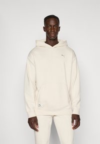 Cremefarvet hættetrøje med en frontlomme, ribbet tekstur og et lille logo. Stoffet er blødt, med matchende tapered bukser for et samlet look.